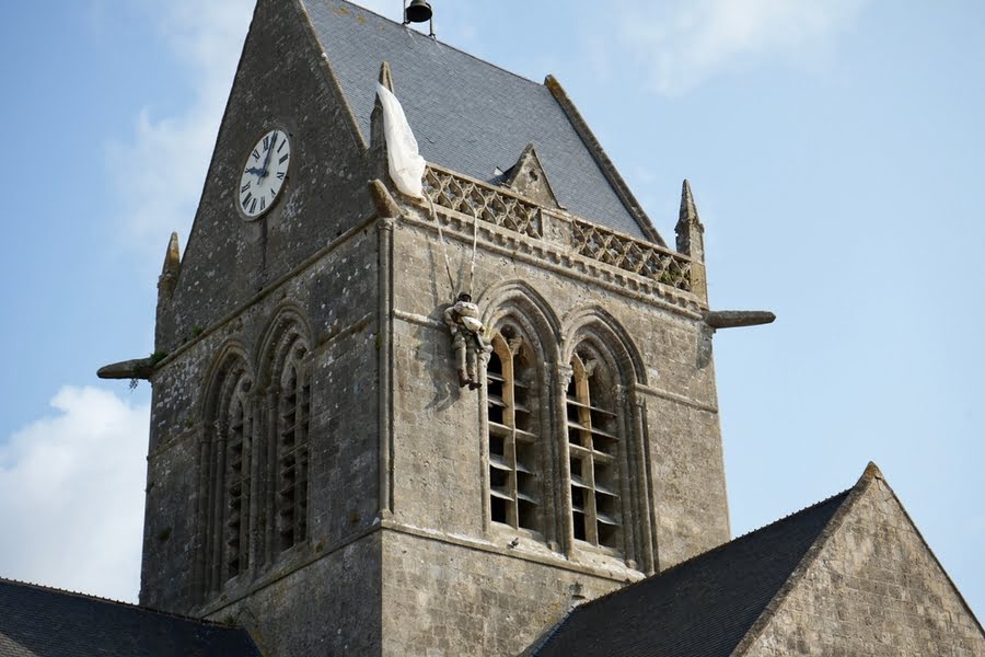 een pop met parachute hangt aan de kerktoren van Sainte-Merè-Église